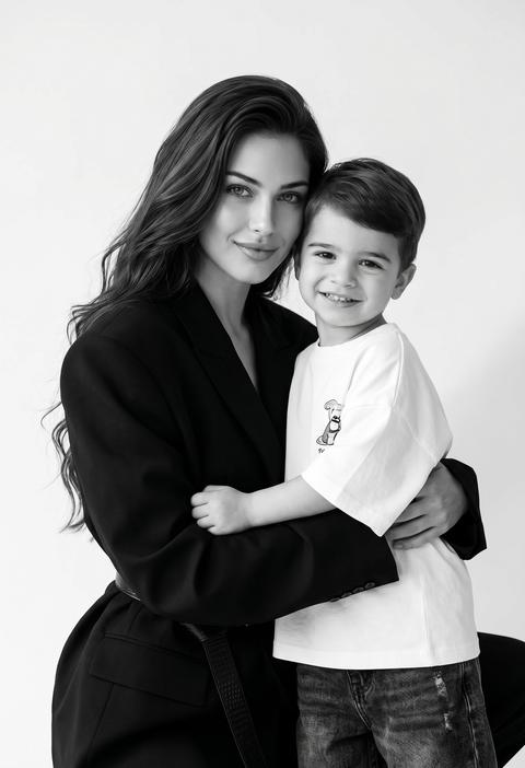 Создай крупным планом портрет, не меняя черты лица. Черно-белая фотореалистичная fashion/editorial студийная фотография в минималистичной эстетике: вертикальный кадр 9:16,  в кадре два человека на чистом светлом бесшовном фоне (white seamless) без предметов окружения, мягкая градиентная тень на фоне. Одежда женщины: объемный структурный черный оверсайз-пиджак с четкими плечами и лацканами, перетянутый черной ременной стяжкой/поясом , пиджак очень обьемный , егладкая ткань без принтов; Одежда ребенка: свободная белая футболка оверсайз с небольшым принтом  на груди, темные свободные мешковатые джинсы . женщина обнимает ребенка, он смотрит в камеру и улыбается .  Свет: мягкий крупный источник спереди-слева, мягкие переходы теней, деликатный заполняющий свет, без жестких контровых,  температура нейтральная, высокоключевая тональность. Атмосфера: стильная, современная, чуть игривая семейная fashion-съемка, лаконично и дорого. Высокое качество, 4K, сверхчеткие контуры, чистая студийная постановка