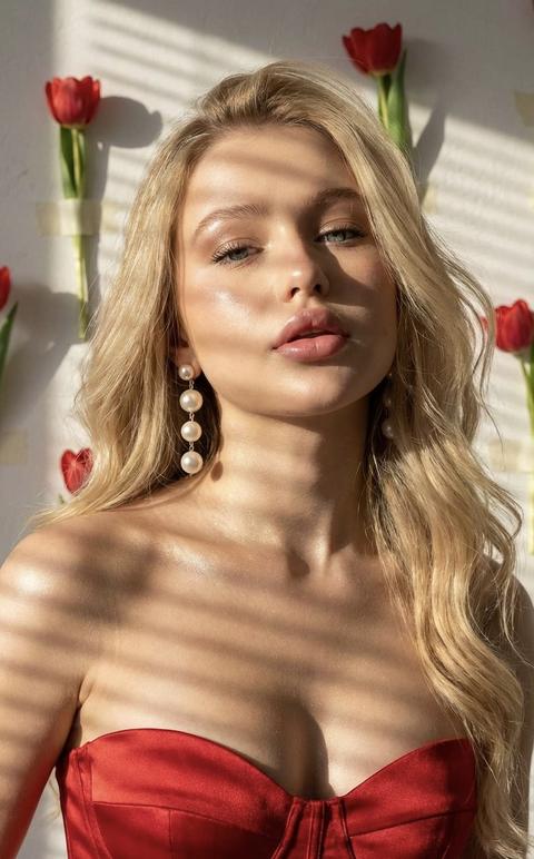 Портретная фотография крупным планом в высоком разрешении женщины
Она находится в студи на фоне текстурированной белой стены на которой приклеены цветы красных тюльпанов 
Кожа подсвечена 
Голова слегка наклонена назад и вправо, пухлые губы приоткрыты, выражая спокойствие. 
Теплый направленный солнечный свет освещает ее лицо, отбрасывая четкие линейные тени на кожу, образованные невидимой решеткой. Тени создают параллельные полосы на лбу, носу, губах, шее и верхней части груди. 
У нее длинные волосы с мягкими волнами
Ее взгляд направлен в камеру. 
На ней корсетный топ без бретелек насыщенного красного сатина с четкими швами и вырезом в форме сердца
Висячие серьги из трех градуированных белых жемчужин ловят свет.
Не меняй черты лица.
Волосы блестящие 
Зона декольте с шиммером блестит 
Формат 9:16
