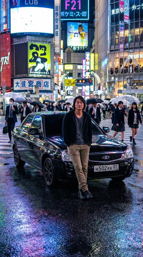 Создать ультра-фотореалистичную уличную сцену в районе Shibuya, Tokyo (Япония) с персонажем Han Lue (Sung Kang) из Fast & Furious, с максимально точной, полностью узнаваемой внешностью 1:1.

👤 ПЕРСОНАЖ:
Han Lue (Sung Kang) стоит перед автомобилем, слегка опираясь на переднюю часть капота.

Внешность строго 1:1:
 • точная форма лица
 • скулы
 • глаза
 • нос
 • губы
 • линия подбородка
 • натуральная текстура кожи
 • возраст
 • фирменная причёска средней длины

Без стилизации
Без сглаживания кожи
Без изменения анатомии
Полный фотореализм

Поза:
 • руки в карманах
 • ноги слегка скрещены
 • расслабленная уверенная стойка
 • взгляд прямо в камеру

Одежда:
 • тёмный худи или куртка
 • серая футболка
 • брюки цвета хаки
 • тёмные ботинки

🚗 АВТОМОБИЛЬ:
Использовать загруженное изображение автомобиля как
ЕДИНСТВЕННЫЙ ТОЧНЫЙ РЕФЕРЕНС (строго 1:1)

Расположение:
 • по центру кадра
 • ракурс: фронт + лёгкий 3/4 (20–30°)
→ чтобы был хорошо виден передний диск

Сохранить:
 • геометрию кузова
 • цвет
 • фары
 • решётку
 • бампер
 • зеркала
 • диски
 • шины
 • стёкла
 • уровень тонировки
 • номерной знак (если есть)
Реалистичный контакт шин с мокрым асфальтом
Физически корректные тени
Естественные отражения неона на кузове и дисках
🏙 ЛОКАЦИЯ — SHIBUYA (НОЧЬ):
Ночной район Shibuya Crossing:
 • огромные LED-экраны
 • японские неоновые вывески
 • вертикальные рекламные панели
 • канадзи и катакана на билбордах
 • светящиеся витрины
 • уличные фонари
 • дорожная разметка перекрёстка
 • мокрый после дождя асфальт
 • отражения неона на дороге
 • стеклянные фасады зданий
 • свет от магазинов и кафе

На фоне:
 • плотный поток пешеходов
 • зонты
 • деловые костюмы
 • школьная форма
 • лёгкое motion blur у людей 

💡 ОСВЕЩЕНИЕ:
Смешанный ночной свет:
 • холодный синий ambient
 • пурпурный и розовый неон
 • зелёные и жёлтые LED-панели
 • тёплый свет витрин
 • верхний городской рассеянный свет

Отражения:
 • на мокром асфальте
 • на лакированном кузове авто
 • на дисках
 • на стекле фар

Контровой свет от билбордов
Лёгкий rim-light по краям силуэта персонажа
Реалистичные мягкие тени

🎥 КАМЕРА:
Вертикальный формат 9:16
Низкий ракурс (уровень бампера)
Cinematic depth of field
Резкий фокус на персонаже и автомобиле
Фон слегка мягкий

🎨 ВИЗУАЛ:
Ultra photorealistic
Cinematic Tokyo night street photography
Wet surface reflections
HDR dynamic range
Sharp focus
Ultra detailed wheel rims
Realistic skin pores
8K

❌ NEGATIVE:
daylight, dry road, front-only angle, hidden wheels, cartoon, CGI, plastic skin, wrong perspective, deformed rims, floating car, fake reflections, low detail, blur
