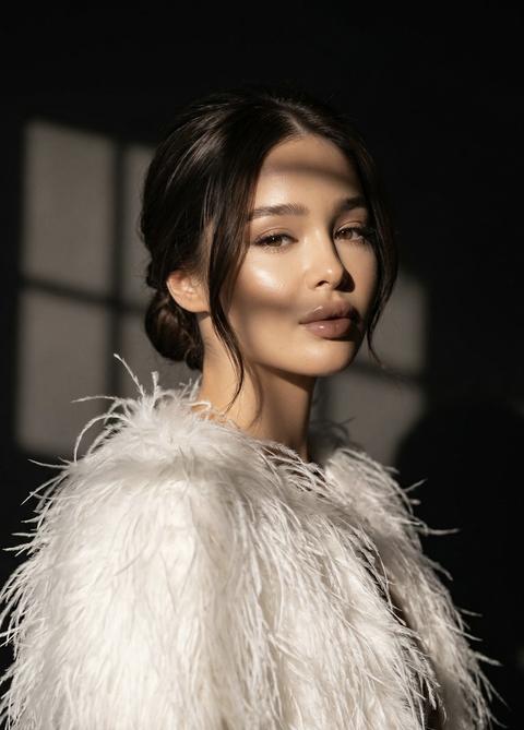 Не меняй черты лица Фотография, женщина в воздушной белоснежной накидке из перьев эффектно позирует в темном студийном помещении с интенсивным боковым освещением и изящно проецируемыми тенями от оконной рамы. Загорелая кожа контрастирует с нежными белоснежными перьями.
Волосы собраны сзади в элегантный низкий пучок, а свободные пряди подчеркивают минималистскую изысканность. Макияж в стиле нюд с глянцевыми губами, подчеркнутыми скулами, нежной подводкой для глаз и объемными ресницами создает утонченный, почти редакторский образ.
Мягкое, объемное освещение создает эффектные формы, Приглушенные теплые тона слоновой кости, шампанского и мягкой бронзы создают неподвластную времени, неземную атмосферу.