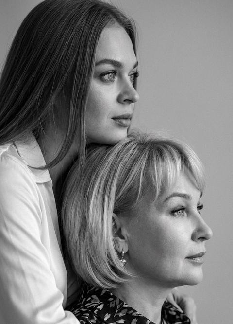 Черно-белый фотореалистичный портрет двух женщин в профиль. В кадре две взрослые женщины, расположенные близко друг к другу, одна позади другой.
Молодая женщина находится сзади и чуть выше, мягко обнимает или опирается на старшую женщину, её подбородок или нижняя часть лица близко к голове старшей. Обе женщины смотрят в одном направлении - вправо, за пределы кадра, с спокойным, задумчивым выражением лица. Молодая женщина, 30 лет с распущеными, естественными волосами. Она одета в светлую рубашку или блузу с длинным рукавом из мягкой ткани, без ярких деталей. Старшая женщина с волосами, аккуратно уложенными. На ней тёмная блуза с мелким узором, возможны небольшие серьги. Одежда повседневная, сдержанная, без броских элементов.
Композиция камерная, крупный план. Кадр обрезан по плечи и голове. Фигуры расположены диагонально, создавая ощущение глубины и близости. Фон однотонный, нейтральный, без деталей, что подчёркивает лица и эмоции. Свет мягкии, направленный сбоку, создаёт плавные тени и подчеркивает черты лица и текстуру кожи. Контраст умеренный, широкий диапазон серых тонов. Стиль классической портретной фотографии, документальный или fine art portrait.
Атмосфера тихая, интимная, эмоционально теплая, с ощущением доверия, поддержки и связи между персонажами. Лёгкое плёночное зерно, высокая детализация, естественные пропорции, отсутствие постановочных поз и искусственной мимики. Размер фото 9:16