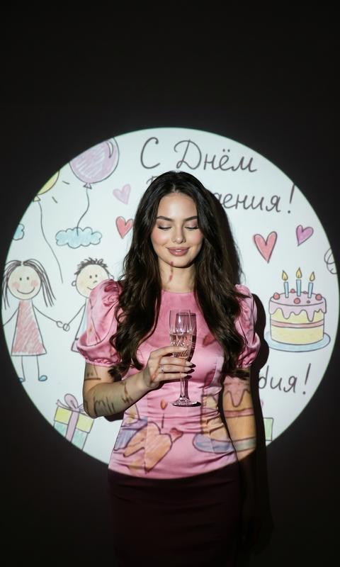 Гиперреалистичный, кинематографический портрет женщины, стоящей немного не по центру внутри кругового луча проектора, снятый от середины бедра до головы. Она свободно держит бокал для шампанского в одной руке на уровне груди, запястье расслаблено, тело тонко наклонено в сторону. Ее глаза мягко закрыты с мягкой, довольной улыбкой, передающей тихую радость и самоанализ.

Ее настоящее лицо остается на 100% неизменным. На ней модное розовое шелковое платье по фигуре с пышными рукавами, аккуратная укладку, естественный макияж и распущенные длинные волосы, мягко обрамляющие ее лицо.

Позади нее нарисованная от руки иллюстрация дня рождения проецируется на стену, заполняя идеальную круглую рамку вокруг ее тела - с детскими рисунками, пастельными цветами, рукописным текстом, сердечками и мультяшным праздничным тортом, надписью с днём рождения .Проекция распространяется по ее одежде и коже, создавая многослойные текстуры и неравномерное падение света.

Освещение низкое и управляемое проектором, с мягкими тенями, приглушенным контрастом и мягкими краями. Фон за пределами проекции исчезает в глубокой темноте, подчеркивая близость и ностальгию. В целом эстетика мечтательна, индивидуальна и кинематографична — как тихий момент дня рождения, запечатленный на пленку, реалистичный и эмоционально теплый.

Настройки камеры:

• Объектив: 35 мм

• Апертура: f/2.8

• Выдержка: 1/80

• ISO: 800

• Освещение: один цифровой проектор в качестве основного источника света

• Фокус: лицо субъекта и верхняя часть торса

• Глубина резкости: от мелкой до умеренной

• Цветовой профиль: мягкие пастельные тона, теплые тона кожи, приглушенный черный

• Текстура: тонкая зернистость пленки, мягкое размытие по краям, естественные недостатки

Отрицательная подсказка:

Иллюстрация, мультфильм, аниме, CGI, 3D-рендеринг, живописный стиль, артефакты искусственного интеллекта, пластиковая кожа, фильтр красоты, чрезмерная резкость, переэкспонированные блики, резкая вспышка, неправильный текст, деформированное тело, лишние конечности, дублирующиеся руки, сильное размытие на объекте, водяной знак, логотип. 3:4