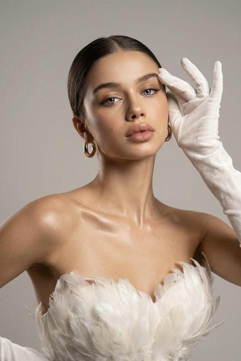 Крупный кадр на объектив 85 мм. Взгляд девушки на камеру. Общее настроение: гламурное, уверенное и немного провокационное — спокойная элегантность с акцентом на линиях плеч и шее. Поза и корпус: корпус слегка повернут в три четверти к камере (плечо ближе к камере смещено вниз), верхняя часть торса чуть наклонена назад, чтобы подчеркнуть линию ключиц. Плечи обнажены, грудная клетка открыта.
Голова и шея: голова слегка наклонена назад и в сторону (туда же, куда повернут корпус), шея удлинена — это создаёт изящную линию от плеч к подбородку. Подбородок чуть приподнят для уверенного выражения.
Руки и жесты: руки в длинных белых бархатных перчатках. Одна рука поднята к голове/виску — пальцы элегантно согнуты, кончики пальцев направлены вверх. Другая рука опущена ближе к корпусу, мягко удерживает край перья на лифе или спокойно лежит на боку.
Одежда и детали: корсет/лиф с большими белыми перьями по линии декольте, создающими объём и текстуру; перья с лёгкой глянцевой/иризирующей поверхностью. Длинные бархатные перчатки — глубокая текстура бархата видна в бликах. Кожа на плечах и ключицах слегка сияющая, с едва заметным блеском для гламурного эффекта.
Серьги золотые кольца. 
• Причёска: гладкие распущенные ,прямо уложенные волосы, откинутые назад вдоль головы. 
• Освещение: мягкий, направленный свет спереди-сбоку (примерно 45°) для мягких теней и рельефа ключиц; небольшой контровой (rim) или боковой свет сзади для отделения силуэта от фона и подчёркивания волос и перьев; слабый заполняющий свет противоположной стороны, чтобы не было слишком глубоких провалов в тенях. На коже — аккуратные горячие блики на ключицах и плечах (легкий глянец).
Фон: нейтральный светло-серый фон с мягкой тенью от фигуры, без лишних деталей, чтобы акцент был на модели и текстурах одежды.
Кадрирование и ракурс: портретный кадр, по вертикали; кадр от середины груди/декольте до чуть выше макушки (плотное, интимное кадрирование). Ракурс на уровне глаз или чуть выше — лёгкий наклон вниз на модель для элегантности.
Глубина резкости и технические параметры: небольшая глубина резкости (портретный объектив 85 мм экв., диафрагма f/2.8–f/4) — фон мягко размытый, объект в фокусе; низкий ISO для чистоты; мягкая обработка с повышенной контрастностью и насыщенностью тёмных тонов, лёгкий виньет для фокусирования на лице и руках.
Постобработка: тёплая тональная коррекция кожи, подчёркнутые блики на ключицах, усиление текстуры перьев и бархата, насыщенные, глубокие чёрные тона, аккуратная ретушь кожи без удаления натуральной текстуры.