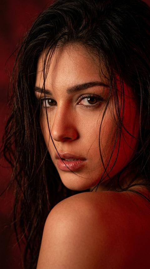 СТРОГО сохранить внешность 1:1 — лицо, черты, пропорции, текстуру кожи, тон кожи, возраст, цвет и длину волос без изменений, без ретуши. Формат 3:4. Фотореалистичный экстремально крупный портрет, кадр очень близкий, лицо занимает почти весь кадр. Камера на уровне глаз. Девушка повернута в 3/4 спиной к камере, через плечо резко оборачивается назад в кадр. Видна верхняя часть открытого плеча на переднем плане снизу справа. Взгляд направлен прямо в камеру, глаза слегка прищурены, выражение напряжённое, чувственное, уверенное. Губы слегка приоткрыты. Волосы распущены, влажные, хаотичные пряди падают на лоб, глаза, щёку и губы, часть волос прилипает к лицу. Свет строго красный, очень насыщенный, low-key, но лицо освещает слабая лампа с теплым светом, фон залит глубоким красным светом без других оттенков. Фон тёмный, почти чёрный с красным свечением. Высокий контраст, живая текстура кожи, влажный блеск на губах