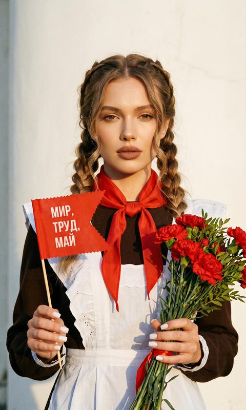 Сделай портрет девушки в стиле «Высокой моды».
Волосы собраны в две толстые косы и волнистые прядки у лица. , 
Маникюр молочный, квадратный , ровный, аккуратный.
Фото в золотой час.
Безупречный макияж, губы пудрово-коричневые. 
На ней форма советских времен , как из СССР, с фартуком 
На шее повязан красный платок на одну сторону, концы платка свободно спускаются вниз, платок короткий.
В одной руке она держит букет красных гвоздик, а в другой флажок красный с вырезанным краем, на флаге написано на четко русском языке по центру: МИР, ТРУД, МАЙ
Теплое, зернистое освещение, высокая контрастность.
Высокая детализация текстур кожи, одежды и волос.
Черты лица полностью сохранены, как на референсном фото.
Фон белая стена.
Без аксессуаров. 