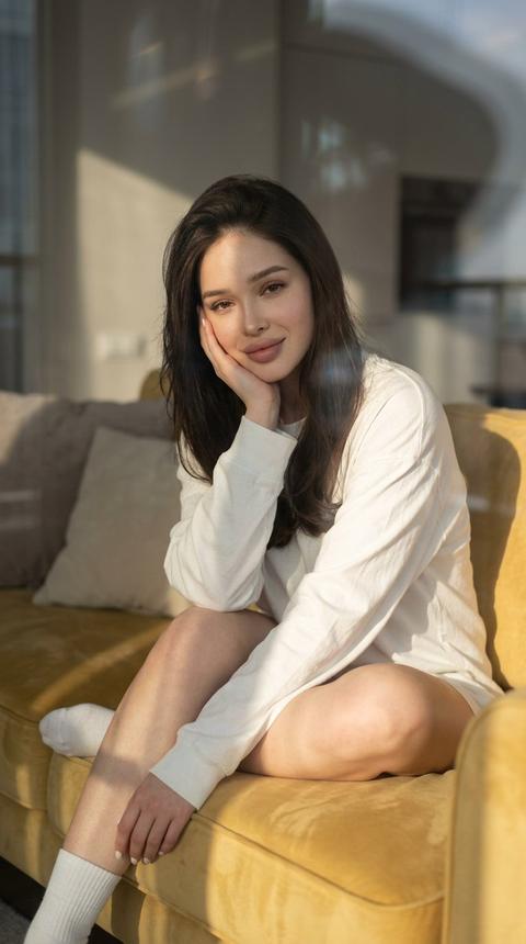 Create a close-up portrait, without altering facial features. An aesthetic, cinematic photo of a young woman sitting in a relaxed pose on a soft yellow sofa in the rays of the setting sun. She is dressed in a cozy white oversized long-sleeve shirt and white socks. The girl gently rests her face on her hand and smiles softly, creating an atmosphere of melancholy and serenity. Flawless makeup, long hair. In the background, soft pillows and a muted interior are visible. Warm golden light (golden hour) falls through the window, creating contrasting shadows and subtle reflections on the glass. High detail in fabric texture, soft focus, depth of field, minimalism and lifestyle photography style.