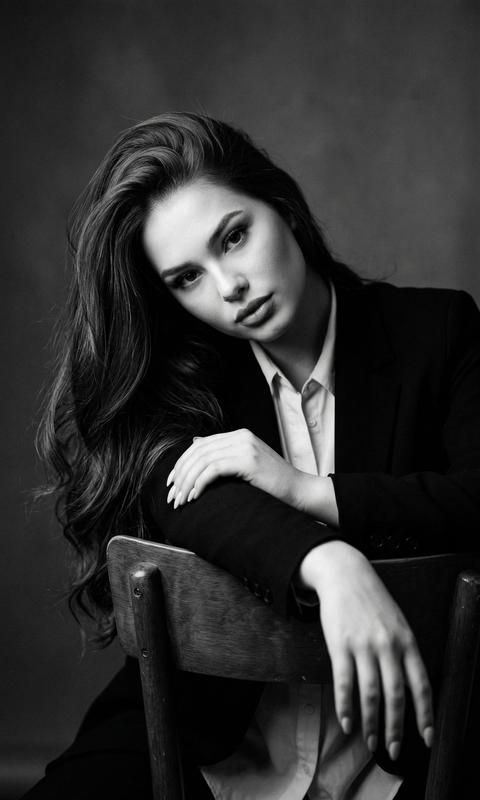 STRICTLY preserve appearance 1:1 — facial features, skin texture, hair color and length. 3:4 format. Black and white studio portrait, chest-up. Camera strictly at eye level, no tilt. Pose strictly: model sits on a chair backwards, torso slightly turned to the right (~20°) and leaning forward. Both hands are draped over the top of the chair back and rest one on top of the other; wrists hang down from the outer side of the backrest. Shoulders relaxed and lowered. Head tilted to the left and forward, chin slightly down. Gaze directed straight into the camera from under the tilt. Hair loose, styled to one side, volume at the roots, partially covering the cheek. Clothing: black jacket, light shirt underneath. The wooden back of the chair is clearly visible in the frame. Dark gray background, softly blurred. Soft side light, deep shadows, high contrast, grain, cinematic, 85mm, f/2.8.