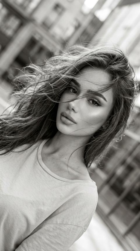 Ультра реалистичное чёрно-белое fashion video frame фото красивой девушки в движении, cinematic monochrome aesthetic, dynamic hair movement, Instagram / Pinterest editorial style, vertical frame 9:16.
Девушка снята крупным портретом под наклоном, будто кадр из видео или случайный стоп-кадр. Камера расположена близко к лицу и немного сбоку, изображение слегка повернуто по диагонали. Девушка смотрит в камеру спокойным притягательным взглядом, губы слегка приоткрыты, выражение лица уверенное, мягкое и немного загадочное.
Волосы распущенные, объёмные, живые. Волосы активно двигаются от ветра или движения головы, часть прядей летит через лицо, глаза, губы и плечи. Пряди должны выглядеть лёгкими, воздушными, немного хаотичными, с натуральной текстурой и мягким блеском. Hair in motion, cinematic movement effect.
Макияж красивый soft glam makeup: ровный сияющий тон кожи, мягкий контуринг, подчёркнутые скулы, деликатный хайлайтер на скулах, носу и над верхней губой. Выразительные глаза, длинные ресницы лисий эффект , аккуратные брови, черная стрелка, мягкие nude shadows. Губы nude glossy lips, объёмные, с мягким контуром и сатиновым блеском. Макияж должен читаться даже в чёрно-белой обработке.
На девушке светлый футболка, мягкая ткань, минималистичный городской образ. Верх одежды слегка открыт у шеи и ключиц, образ элегантный, не перегруженный.
Свет мягкий дневной, natural city daylight, без вспышки. Свет идёт сбоку и сверху, подсвечивает лицо, волосы, губы и ключицы. На волосах светлые highlights, на коже мягкое сияние. Тени плавные, без жёсткой студийности. Свет выглядит как естественная съёмка на телефон на улице или возле городской архитектуры.
Цветокоррекция чёрно-белая, soft monochrome grading, мягкие серые полутона, светлые highlights на волосах, глубокие, но не проваленные тени, low contrast editorial look, slight blur from movement, subtle film grain. Кадр должен выглядеть живым, немного размытым, как стильный стоп-кадр из Reels.
Фон городской, сильно размытый: архитектура, улица, фасады зданий, стекло или витрины. Background blur, shallow depth of field, город не должен отвлекать, главный акцент на лице, волосах и движении.
Композиция: close-up / medium close-up shot, diagonal tilted frame, лицо девушки занимает центр кадра, волосы пересекают кадр динамичными линиями. Вертикальный формат 9:16, ощущение движения, лёгкая неидеальная резкость, aesthetic mobile video frame.
Качество: снято на iPhone 17 Pro, ultra realistic mobile photography, cinematic black and white video frame, realistic skin texture, detailed moving hair strands, natural motion blur, soft focus, 4K, без мультяшности, без plastic skin.
Negative prompt:
color photo, perfectly still hair, slick bun, harsh flash, studio background, looking away, wide smile, teeth showing, overexposed face, messy low quality blur, plastic skin, cartoon face, CGI look, distorted eyes, distorted face, bad anatomy, extra fingers, low quality.