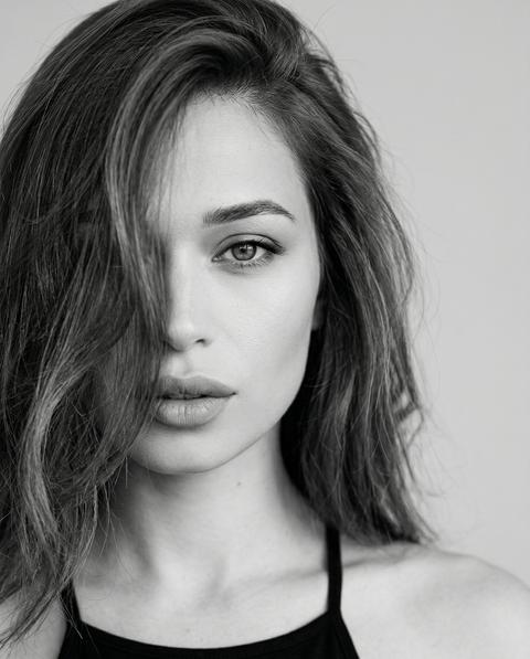 СТРОГО сохранить внешность 1:1 — лицо, черты, пропорции, текстуру кожи без изменений. Формат 3:4. Чёрно-белый фотореалистичный экстремально крупный beauty-портрет на светлом фоне. Камера строго на уровне лица, объектив параллелен полу. Кадрирование асимметричное и очень плотное: левая часть лица сильно срезана левым краем кадра, левый висок, часть лба, часть левой щеки и край губ вне кадра. Лицо смещено влево, не по центру. В кадре полностью виден только правый глаз, правая бровь, правая щека, нос, правая часть губ, подбородок, шея, левая часть лица и волос выходит частично за пределы кадра. Голова слегка опущена вниз. Взгляд томный, уверенный, строго в камеру. Губы слегка приоткрыты. Волосы распущены, объёмные, густо закрывают левую сторону лица и частично левый глаз, несколько прядей спадают на лоб. Одежда: чёрный топ с тонкими бретелями. Мягкий контрастный black and white, clean background, фотореализм