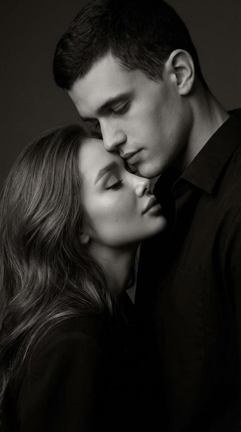 Создай портрет, не меняя черты лица. Кинематографический монохромный снимок крупным планом - женщина слегка наклоняется, ее лицо обращено к склонившемуся над ней мужчине, его голова слегка приподнята над бровями.
Их глаза слегка прикрыты.. у женщины длинные волнистые волосы, безупречный макияж. 
Их лица сближены. Его подбородок касается ее лба.
Освещение: низкий софтбокс с матовой отделкой в тон; оттенки черного плавно переходят в серый, чтобы подчеркнуть складки и переплетение ткани.
Тонкое свечение в полутонах, реалистичные детали кожи, органическая текстура.
Фон плавно переходит в виньетку, без резких контрастов.
Настроение: мягкая связь, художественная близость, изысканно-черная редакция
