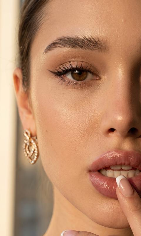 Сохранить внешность 1:1. Фото-единственный эталон. Экстремальный крупный план: правая половина лица, фокус на глазе и губах. Взгляд глубокий, cat-eye стрелки, длинные ресницы. Голова совсем слегка наклонена. Фон светлый, дневной свет. Губы нюд «пыльная роза», глянец, слегка приоткрыты; палец с френч-маникюром у губ. Свет: тёплый боковой (golden hour), мягкие тени, блики на коже и серьгах. Камера: 85mm, f/1.4, кремовое боке, детализация кожи.
