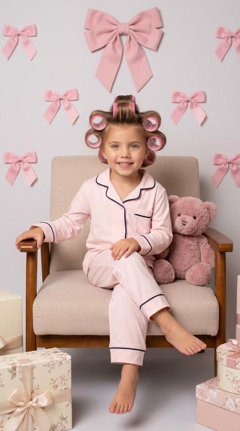 A little girl sits in a soft chair with wooden armrests. Her body is upright, her legs are neatly crossed, her left hand is on the armrest, her right hand is on her knee, and she is looking directly at the camera. The shot is medium-wide, from the feet to the top of her head, the angle is at the child's eye level. The composition is centered, the figure is positioned precisely in the center, and the surrounding space is symmetrically organized.

Her hair is styled in large pink curlers evenly distributed over her head, the surface smooth and neatly divided into strands.

She is wearing light pink pastel pajamas made of soft cotton with a matte finish. Dark blue contrasting piping is trimmed around the collar and cuffs. The fabric is smooth with slight natural folds. The pajamas have long sleeves and long pants. The chair is upholstered in a light beige fabric with a fine texture, and the wood elements are a warm brown with a matte finish. To the right, a muted dusty pink teddy bear with a soft, fuzzy texture sits in a chair. Gift boxes in cream and light pink shades with a floral print and pink satin ribbons are placed on the floor. On the wall behind her is a light gray backdrop with evenly spaced decorative pink bows and a large, airy dusty pink bow at the top of the frame.

The lighting is soft studio lighting, with the main diffused light positioned frontally and slightly above eye level. The light is neutral to warm, evenly illuminating the face and objects. The shadows are soft and smooth, the contrast is low, and the background is evenly lit without pronounced gradients.

Shot with a 50mm lens, moderate depth of field with sharp focus on the face, slightly softened background, and high texture detail in the fabric, wood, and plush. Staged studio portrait photography, highly detailed, photorealistic.

soft pastel studio mood,
dominant light pink and creamy beige tones,
neutral-warm color temperature,
low to moderate saturation with pastel emphasis,
low contrast with soft shadow transitions,
diffused frontal lighting,
gentle highlight reflections,
natural soft skin tone rendering,
clean smooth texture with minimal grain,
bright pastel color grading. Не меняй черты лица 