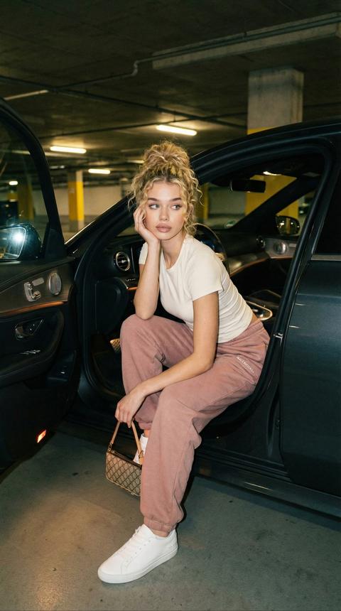 Ночной портрет у автомобиля в полутёмном паркинге, вертикальный кадр от середины бёдер. Женщина сидит на краю открытой двери, корпус слегка наклонён вперёд, в руке — маленькая дизайнерская сумка, взгляд в сторону, спокойное выражение лица.
На ней облегающая айвори-футболка и пудрово-розовые брендовые джоггеры свободного кроя, минималистичная обувь. Волосы кучерявые  собраны в небрежный высокий пучок, украшения минимальные. Приглушённый свет из салона авто и ламп паркинга, лёгкая вспышка смартфона. Натуральный макияж, плёночное зерно, clean girl × luxury lifestyle, фотореализм, 8K