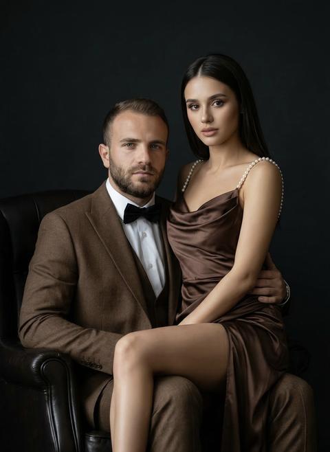 Не меняя внешности людей с фото, сцена погружена в приглушённый, глубокий тёмный фон, создающий атмосферу интимности и элегантности, где каждая деталь подсвечена мягким боковым светом, акцентируя линии тела и фактуру одежды. Первый мужчина сидит в кресле, строго и уверенно, в классическом коричневом костюме с чётко отглаженными складками и белой рубашкой под ним, украшенной аккуратным чёрным бабочкой. Его осанка прямая, руки расслаблены — одна обвивает талию женщины, другая скрыта тенью, создавая ощущение поддержки и гармонии между ними. Лицо мужчины освещено равномерно, подчёркивая рельефность черт, взгляд направлен вперёд с лёгкой степенью задумчивости и спокойствия.  Первая женщина сидит на коленях мужчины, корпус её слегка развернут в сторону камеры, придавая позе плавный и изящный изгиб. Она облачена в блестящее шоколадное платье из атласа с драпированной лифом, которое эффектно отражает свет, подчёркивая изящество линий тела и мягкость текстуры ткани. Лямки платья украшены жемчужными бусинами, хорошо различимыми благодаря аккуратному световому контрасту. Платье длинное, с разрезом, обнажает одну ногу полностью — гладкую, стройную, на ногах аккуратные бежевые босоножки на тонких ремешках, которые красиво переплетаются вокруг щиколоток. Волосы женщины ниспадают темными волнами через плечи, создавая выразительную рамку для лица с естественными чертами и сосредоточенным взглядом, направленным прямо в камеру, что придаёт портрету сильное эмоциональное присутствие.  Общая композиция выдержана в стиле классической гламурной фотографии с акцентом на контраст между холодной строгостью костюма и мягкой текучестью атласного платья. Свет и тени тщательно играют роль в создании объёма и глубины, усиливая ощущение роскоши и близости между персонажами, их гармоничной взаимосвязи. Тёмный нейтральный фон не отвлекает внимание, а напротив — подчёркивает тонкость нюансов освещения и фактуры тканей, создавая эффект трёхмерности и реалистичного присутствия в кадре, фотореалистичное изображение, высокая текстурная детализация, высокое качество, разрешение 8K.