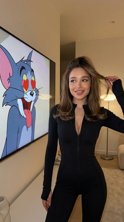 Сохранить внешность 1:1. Не изменять черты лица, пропорции. Игривый и яркий портрет девушки. Кожа сияет, естественный макияж. Она позирует в уютном минималистическом современном интерьере. Она одета в облегающий черный спортивный комбинезон, слегка растегнутый. Девушка стоит корпусом к камере, смотрит в сторону телевизора, живое фото, рука поправляет волосы в движении, у нее красивая укладка закрученными к низу волосами с легким объемом у корней. На заднем плане виден очень большой ЖК-экран, на котором замер кадр из мультфильма «Том и Джерри» с котом Томом, у которого вместо глаз красные сердечки и высунутый язык. Без надписей. Мягкое домашнее освещение создающее эффект живого фото из соцсетей, минималистичный декор квартиры. Высокое разрешение, реалистичные текстуры кожи и ткани. Выдержка фото понижена