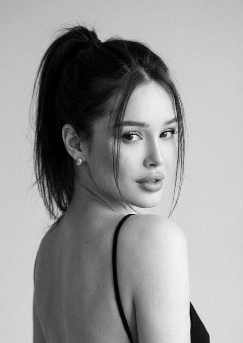 СТРОГО сохранить внешность 1:1 — лицо, черты, пропорции, форму глаз и губ, текстуру кожи, длину и густоту волос; фото — единственный эталон. Без ретуши, без сглаживания, без изменения анатомии. Чёрно-белый фотореалистичный портрет, вертикальный формат 3:4, tight medium close-up от середины спины и выше. Девушка стоит спиной к камере, корпус развернут примерно на 60° влево, открытая линия спины. Голова повернута через плечо к камере, взгляд прямой, спокойный, немного томный. Губы слегка приоткрыты, выражение уверенное и мягкое. Волосы собраны в высокий хвост, несколько тонких прядей свободно падают на лоб и щёки. Одежда: чёрный минималистичный топ на тонких бретелях, глубокий вырез по спине. Украшение — небольшие гладкие серьги без блеска. Свет мягкий рассеянный спереди и чуть сверху, низкий контраст, плавный градиент теней, лёгкое плёночное зерно. Фон светло-серый студийный, без текстуры. 85mm, f/2.0, фокус строго на глазах, гиперреализм, 8K. СТРОГО сохранить внешность 1:1 — лицо, черты, пропорции, форму глаз и губ, текстуру кожи, длину и густоту волос; фото — единственный эталон. Без ретуши, без сглаживания, без изменения анатомии. Чёрно-белый фотореалистичный портрет, вертикальный формат 3:4, tight medium close-up от середины спины и выше. Девушка стоит спиной к камере, корпус развернут примерно на 60° влево, открытая линия спины. Голова повернута через плечо к камере, взгляд прямой, спокойный, немного томный. Губы слегка приоткрыты, выражение уверенное и мягкое. Волосы собраны в высокий хвост, несколько тонких прядей свободно падают на лоб и щёки. Одежда: чёрный минималистичный топ на тонких бретелях, глубокий вырез по спине. Украшение — небольшие гладкие серьги без блеска. Свет мягкий рассеянный спереди и чуть сверху, низкий контраст, плавный градиент теней, лёгкое плёночное зерно. Фон светло-серый студийный, без текстуры. 85mm, f/2.0, фокус строго на глазах, гиперреализм, 8K.