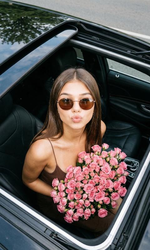 Девушка сидит в дорогой машине черной с люком 
Фото сделано сверху в открытый люк 
Девушка подняла голову вверх на камеру 
Она одета в коричневый топ на тонких бретелях. Очки круглые с коричневыми линзами 
На коленях у нее огромный большой букет кустовых розовых роз
Волосы распущенные  блестящие 
Она смотрит в камеру губами делает поцелуй 
Формат 9:16 
Освещение дневное с легкой зернистостью 
Кинематографический эффект