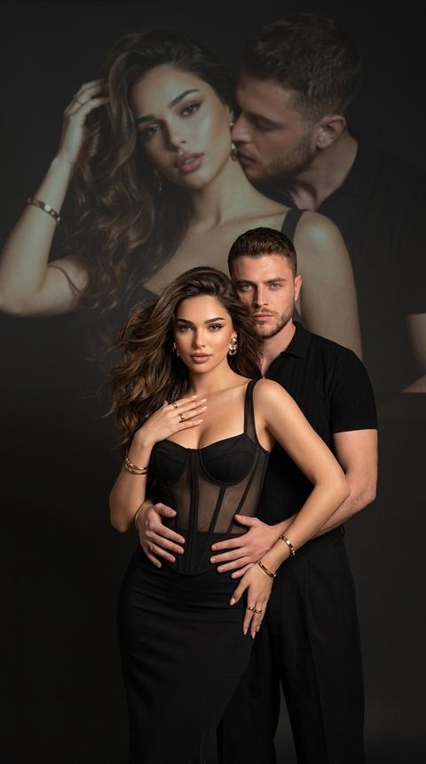 Создай , не меняя черты лица. фотография мужчины и 
женщины в элегантной позе. Она одета в обтягиваютый корсет с прозрачными элементами и длинную, облегающую черную юбку. Ее  объемные волнистые волосы, частично закрывая ее лицо, мужчина стоит немного позади обнимает ее за талию, смотрит в камеру. Одна из ее рук касается области груди, в то время как другая слегка находится на бедрах, что создает динамичную позу. Она выглядит уверенно и привлекательно. Она носит массивные золотые серьги. На руках золотые браслеты и кольца . Мужчина в черной рубашке и свободных брюках. Они покрывает меньшую часть
Фон темный и минималистичный, в верхней части изображения есть мягкая и тусклая проекция
одной и той же пары (в другой позе).
Естественная цветовая проекция мужчины и женщины занимает большую часть верхней части
изображения и выглядит значительно больше. У нее соблазнительный взгляд,  держа  волосы одной рукой, и соблазнительно смотрит. Он целует ее в шею. Мужчина выше женщины. Максимальная / детализация 