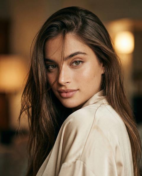 СТРОГО сохранить внешность 1:1 — лицо, черты, пропорции, текстуру кожи, фото — эталон внешности. Формат 3:4. Кинематографичный бьюти-портрет по плечи. Модель в драматичной over-the-shoulder позе: корпус слегка отвернут от камеры, голова повернута к объективу. Взгляд в камеру, мягкая уверенная улыбка. Волосы распущены, свободно падают на плечи, боковой пробор, несколько прядей частично закрывают лицо, создавая модный editorial-эффект. Одежда: лёгкий кремово-бежевый сатиновый топ с мягкой драпировкой на плечах. Макияж: мягко подчеркнутые скулы, длинные ресницы, лёгкий лайнер, губы приглушённый розово-нюд. Фон минимальный, тёплые нейтральные оттенки, мягко размытый. Свет тёплый контрастный боковой, создаёт объём лица. Съёмка на ретро 35mm плёночную камеру, объектив 50mm, малая глубина резкости. Тёплый винтажный цвет, заметное плёночное зерно, лёгкий glow, ощущение старой fashion-съёмки, высокая детализация, фотореализм.