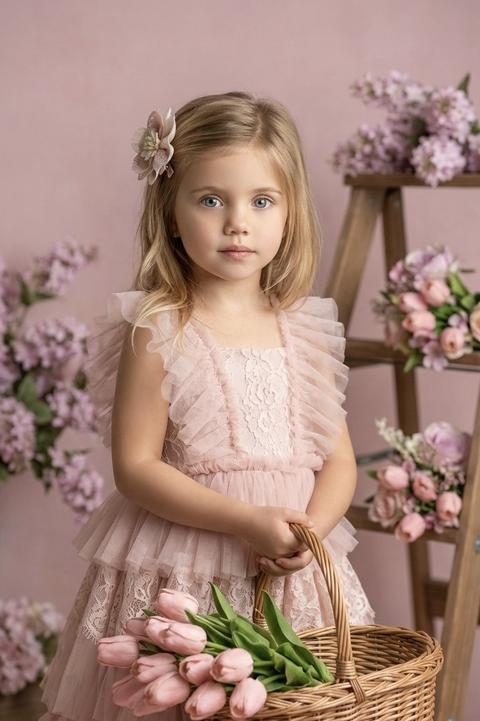 A little girl stands in a studio setting with floral arrangements, her body turned toward the camera, holding a wicker basket with a large bouquet of tulips at waist level with both hands. Her elbows are slightly bent, her shoulders are relaxed, and her head is held straight, looking directly at the lens. The medium shot extends from her hips to her head, the angle is at the child's eye level. The composition is centered, emphasizing the basket and the volume of the flowers, while the background gently recedes into the distance.

Her hair is pinned to the side with a decorative fabric flower clip.

She is wearing a powder-pink dress made of multi-layered, translucent fabric with lace inserts and ruffles on the sleeves and hem. The bodice has a textured trim, and the skirt is full and layered, the fabric is lightweight and matte. She holds a wicker basket made of natural material with a pronounced weave, inside which is a dense bouquet of tulips with closed, soft-pink buds and green leaves.

The backdrop is a decorative wall in a soft pink hue, with lilac-pink floral arrangements and a wooden stepladder with bouquets displayed in the background. The space is designed as a studio flower garden.

The lighting is soft studio lighting, with the main light positioned in front and slightly to the side. The light is diffused and even, creating soft shadows under the chin and along the arms. The contrast is moderate, the transitions between light and shadow are smooth, the highlights on the petals and fabric are delicate and controlled, and the background is softly lit without sharp brightness changes.

Shot with an 85mm portrait lens, shallow depth of field with sharp focus on the face and the top of the basket, the background is softly blurred, and the skin texture, fabric, basket weave, and petals are highly detailed. This is a highly detailed, photorealistic children's art portrait.

soft romantic studio mood,
neutral to slightly cool color temperature,
moderate saturation with pastel emphasis,
medium-low contrast with smooth shadow transitions,
diffused soft lighting,
natural soft skin tones,
gentle highlight roll-off,
smooth background blur with depth,
floral editorial color grading. Не меняй черты лица 