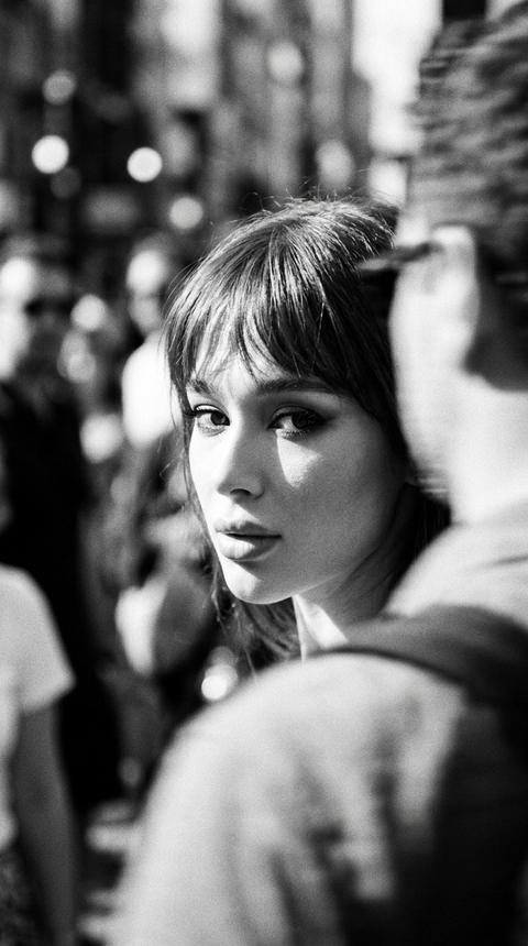 Cinematic effect with added grain, black and white photo.
Loose hair with bangs, in Old Money style.
Extreme close-up of face, no visible clothing. Sun partially illuminates the face.
Bright makeup. A girl stands in a crowd and looks over her shoulder into the camera. The image is intentionally blurred and out of focus, with noticeable motion blur, creating a dreamlike and impressionistic feel, as if it were a captured memory.
People around are motion blurred and smudged.
Face is clearly visible.
Do not change facial features.
Format 9:16.