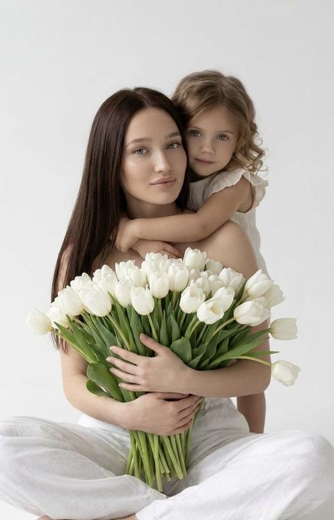 Создай крупный  фотопортрет , не меняя черты лица, молодой девушки с  ухоженными волосами, сидящей в позе лотоса на белом фоне. Безупречный нежный макияж. Она держит в руках огромную охапку белых тюльпанов, которые закрывают часть её тела. На ней свободные белые брюки.сзади её обнимает девочка, на девочке светлый топ с рюшами, волосы с легки локонами.  Минималистичный стиль, мягкое естественное освещение, пастельные тона, высокая детализация текстуры кожи и ткани. Эстетика чистоты, нежности и весны. Белый студийный фон, мягкий рассеянный свет, кинематографичное качество, 8k, фотореализм.