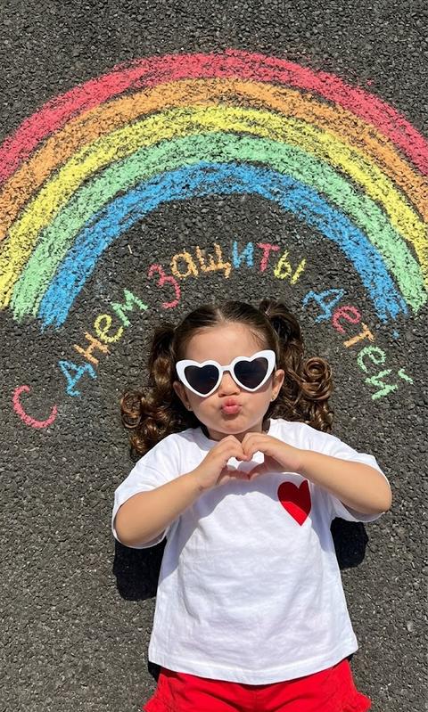 create without changing facial features
Top-down shot of a small child lying on dark asphalt, in the center, with a bright, chalk-drawn rainbow arching over their head. The child is wearing white heart-shaped glasses. White oversized t-shirt with a red heart on the right side. Red ruffled shorts. Hands in a 🫶🏻 gesture. Lips pursed for a kiss. The chalk rainbow has distinct stripes of red, orange, yellow, green, and blue with a slightly faded texture. The asphalt has a rough, speckled texture. Bright, clear daylight casts distinct, sharp shadows from the child onto the asphalt, indicating a high sun angle. The overall mood is playful and summery. On top of the asphalt, written in colorful chalk, is "Happy Children's Day". Hair with curls at the ends.