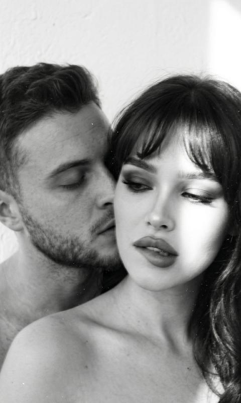 Кинематографический эффект с добавлением зерна черное белое фото
Волосы у девушки распущенные с челкой, в стиле Олд Мани 
Экстремально крупный план лиц, без видимой одежды. Она кусает нижнюю губу не смотря в камеру За ней скос на целует её в шею с закрытыми глазами  Солнце частично освещает лицо 
Макияж у девушки яркий Они стоят на фоне белой стены. Изображение намеренно размыто и имеет расфокусировку, с заметным размытием движения, создавая ощущение сновидения и импрессионизма, словно это запечатленное воспоминание.
Лица четко видны 
Не меняй черты лица
Формат 9:16