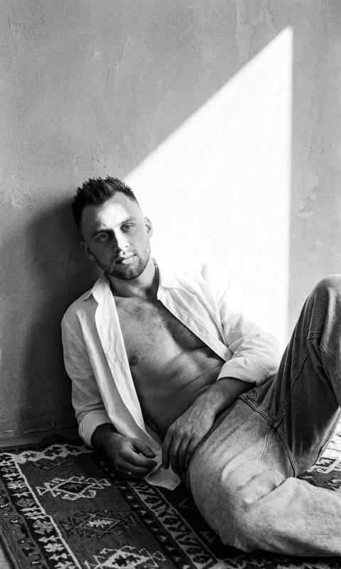 Создай крупный портрет, не меняя черты лица. Кинематографический эффект с использованием зернистости. Черно-белый снимок мужчины, который лежит на полу на ковре с этническим арнаментом, прислонившись к стене в расслабленной позе. Он смотрит в камеру. На нем свободные джинсы и белая  рубашка, пуговицы расстегнуты на рубашке. мужчина спортивного телосложения,  Яркий солнечный свет падает на стену  полосой света. Он задумчиво смотрит в камеру,  Формат 9:16