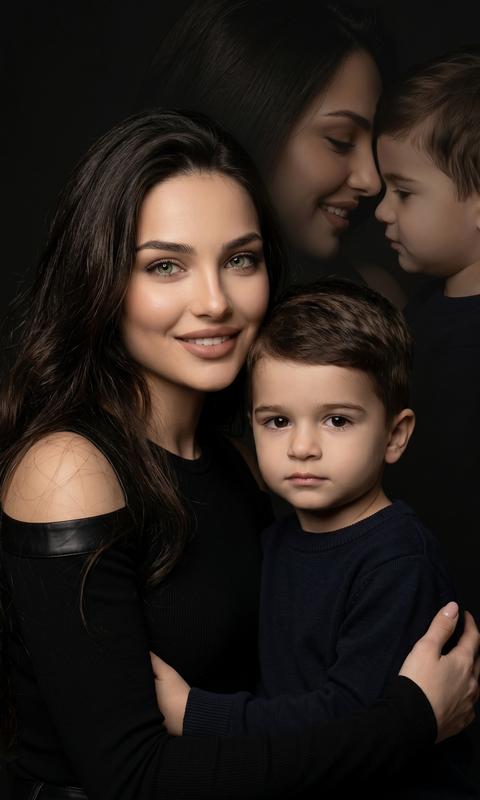 Create a large portrait, without changing facial features, transfer exactly from the attached photo. Colorful cinematic expressive studio close-up portrait.
Close-up focused on the faces of a woman and a child.
The woman stands behind, embracing the child, looking tenderly at the camera. She looks at the camera, her makeup is flawless, lips powdery brown, hair long and wavy. She is wearing a black off-the-shoulder long-sleeve top.
The boy is positioned in the foreground, dressed in dark clothing. In the background, a double exposure, barely visible silhouettes of the same woman and child, a close-up shot, their faces in profile looking at each other with tenderness and smiles. The background is dark. The light is soft but dramatic, highlighting only the facial features. High resolution, cinematic effect, high detail of skin, hair, clothing, 8k.