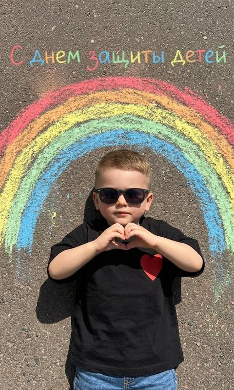 create without altering facial features
Top-down shot of a small child lying on dark asphalt, in the center, and above their head, a bright, chalk-drawn rainbow arches. The child is wearing dark glasses. A black oversized t-shirt with a red heart on the right side. Denim shorts. Hands in the 🫶🏻 gesture. The chalk rainbow has distinct stripes of red, orange, yellow, green, and blue colors with a slightly faded texture. The asphalt has a rough, mottled texture. Bright, clear daylight casts distinct, sharp shadows from the child onto the asphalt, indicating a high sun angle. The overall mood is playful and summery. On the asphalt, written in colorful chalk from above, "Happy Children's Day"
