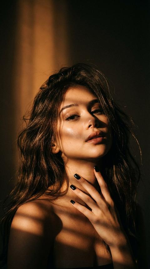 Низко-киновая портретная композиция в
вертикальном формате, кадр захватывает шею,
плечи и верхнюю часть торса.
Много тёмного
негативного пространства вокруг.
Тёплый направленный свет (оранжево-золотистый) , падающий под углом сверху/слева; свет проходит через узкую решётку/жалюзи и отбрасывает чёткие параллельные полосы теней по хоже плеч и груди.
Высокий контраст: глубокие чёрные тени и яркие,
HO
тёплые световые блики на коже;
мягкие
отражения на боковых прядях волос. Длинные очень
объемные растрепанные волосы, глянцевые,
тонкими бликами на отдельных прядях; волосы
свободно спадают по бокам, создавая текстурный
обрамляющий фон. серный квадратный маникюр,
Поза
девушки расслабленная , подбородок немного задрат,
смотрит в камеру. одной рукой держится элегантно за шею , губы слегка приоткрыты. Кожа тёплого оттенка, с видимой текстурой и натуральными бликами; линии тени подчёркивают форму ключиц и контура шеи. Общая атмосфера - драматичная,
интимная, лёгкая загадочность;
кинематографичный, артхаусный стиль съёмки. Цветовая палитра: глубокие тёплые коричневые и золотистые тона на светах, насьщенные почти чёрные тени, минимальная насыщенность фоновых цветов. Резкость в зоне света на
снижение резкости по
краям;
мелкое зерно или лёгкая плёночная
текстура для реалистичности. Возможные технические параметры промта: низко-ключевой
HDR, фокус на техстуре кожи и волос, shallow depth of field (широкая апертура), точечный боковой источник света через полосатую маску/ жалюзи,
50-85 mm,
cinematic lighting, dramatic shadows.
Не меняй черты лица