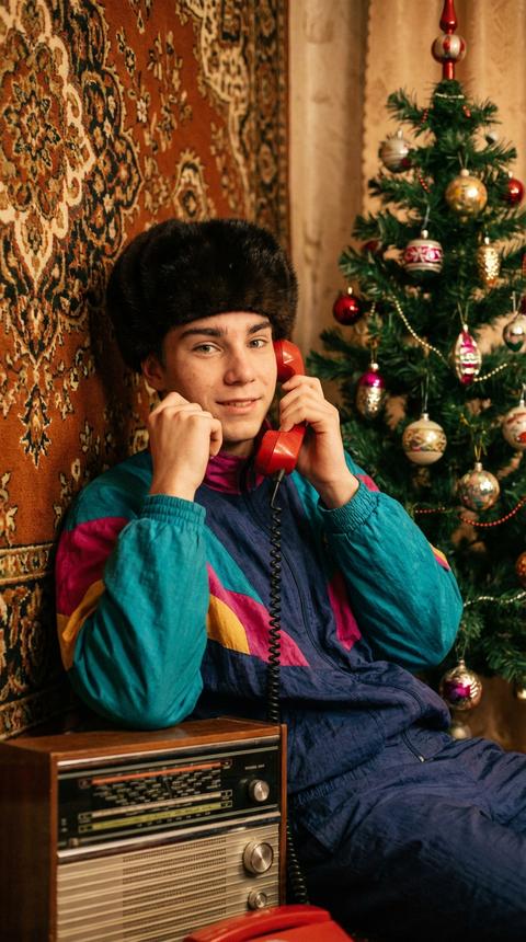 На фотографии изображен мужчина в ретро-стиле, который создает атмосферу новогодней фотосессии. Он одет в спортивный костюм в стиле 90х дополненный меховой норковой шапкой, что придает образу праздничность. В руках у него красный телефон с длинным шнуром, который является ярким акцентом и символизирует эпоху 80-х годов. Фон фотографии украшен ковром с богатым орнаментом, что добавляет интерьеру винтажный шарм. Справа видна часть новогодней елки, украшенной игрушками, что подчеркивает праздничное настроение. На переднем плане стоит радиоприемник, который усиливает атмосферу ретро. Поза мужчины расслабленная, но элегантная: он слегка опирается на стену, держа телефон у уха, что создает ощущение непринужденности и уюта. Общий стиль фотографии передает дух 80-х годов с элементами новогоднего настроения. Он смотрит прямо в кадр, сорани исходные черты лица, профессиональная частота резкости 85mm,