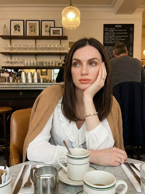 Фотография женщины, сидящей за столиком кафе, она опирается щекой на левую руку и смотрит немного вверх и в сторону с задумчивым выражением лица, поясной кадр, центрированная композиция с женщиной в центре и интерьером кафе, заполняющим фон. Кожа ровного матового тона, веки с теплыми коричневыми тенями, ресницы длинные и подчеркнутые тушью, губы с помадой персиково-нюдового оттенка. На ней свободная белая блузка с V-образным вырезом и отделкой рюшами, вязаный кардиган верблюжьего цвета наброшен на ее плечи, рукава длинные и слегка свободные, тонкий золотой браслет и браслет из белого жемчуга на ее левом запястье, и простое золотое кольцо на правой руке. Перед ней на мраморном столике кафе стоят несколько белых керамических кружек с зелеными полосками, одна с металлической ложкой внутри...