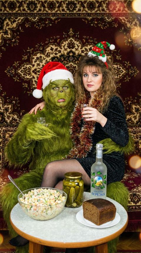 На фотографии изображена необычная и юмористическая сцена. Фотография передает дух русского нового года в стиле 90×. 
В центре композиции — два персонажа, сидящих рядом на фоне роскошного бордового ковра с восточным орнаментом. Слева пьяный Гринч из фильма  похититель Рождества  в красной шапке Санты с белым помпоном. Гринч держит в руке рюмку водки, его выражение лица кажется слегка удивлённым и пьяным.Справа от него — девушка в блестящем черном платье, немного вульгарном на шее висит елочноя мишура. На голове у нее смешной  новогодний колпак и пышные локоны.  Она  тепло обнимает Гринча  за плечо сидя у него на коленях  в другой руке держит рюмку водки. Они смотрят в кадр.
Перед ними расстоложен круглый стол с русскими закусками. 
большая миска с салатом (похоже на оливье или аналогичный праздничный салат); 
банка маринованных огурцов; 
бутылка водки «Березка» с узнаваемой этикеткой; 
кусок чёрного хлеба. 
На фоне коврана стене. 
Композиция выглядит как ироничная интерпретация традиционного семейного застолья, где вместо обычного гостя — знаменитый персонаж из западной сказки. Атмосфера одновременно уютная и комичная, благодаря сочетанию контрастных элементов: сказочного героя, народных мотивов и классических закусок. Фон с ковром добавляет сцене «домашнего» уюта и подчёркивает колорит. 
Не меняй исходные черты лица девушки, сохрани исходные черты лица, в кадре присутствует легкое боке подчеркивающее юмористическую ситуацию, фото со вспышкой, запрещено мультяшное изображение.