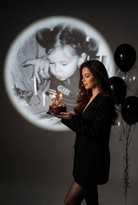 Используйте внешность пользователя в масштабе
1:1 сте же лицо, тон кожи, волосы и пропорции то без каких-либо изменений.
Фотореалистичное художественное фото на день рождения в тёмной студии. Пространство минималистичное, с мягким рассеянным светом и глубокими тенями. Девушка стоит в профиль, слегка опустив взгляд, в руках держит небольшой торт с зажженными свечами в виде цифр {age}. Тёплый свет свечей деликатно подсвечивает лицо и ладони, создавая интимное ощущение момента.
На фоне - большая круглая проекция чёрно-белой детской фотографии самой девушки, с проектора.
Детский портрет слегка размытый по краям, с мягким световым ореолом, как воспоминание или образ из прошлого. Черты ребенка узнаваемы, но мягким световым ореолом, как воспоминание или образ из прошлого. Черты ребёнка узнаваемы, но не слишком резкие, чтобы сохранить ощущение ностальгии и времени. Образ взрослой девушки пО сдержанный и элегантный: чёрный удлинённый жакет или лаконичное платье,/чёрные ИИ полупрозрачные чулки, минималистичный силуэт.
Волосы распущены, естественные, без сложной укладки. Макияж нейтральный, с акцентом на ровную кожу и спокойный, осознанный взгляд.
Справа в кадре - связка чёрных и прозрачных воздушных шаров, добавляющих символ праздника, но не отвлекающих от основного сюжета. Глубина резкости мягкая, фон слегка размытый. Кадр вертикальный, кинематографичный, с лёгкой зернистостью.
Настроение - ностальгия, принятие, взросление, день рождения как личная точка отсчета.