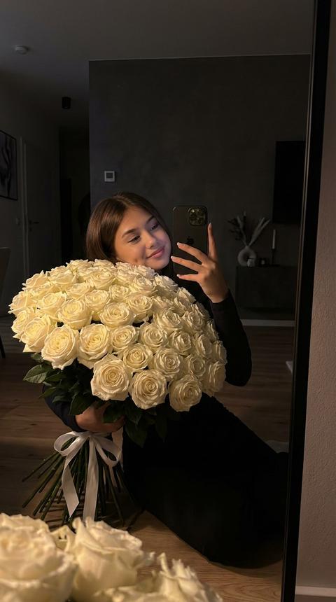 Реалистичное фото девушки, в 8к качестве , где она себя фотографирует на телефон в зеркало, где в центре внимания находится роскошный букет из большого 301 белой розы, которые полностью лежат на полу. Девушка сидит на полу, и немного касается букета. Букет заполняет основную часть кадра, нижняя часть кадра полностью заполнена букетом. его лепестки блестят на свету, подчеркивая их яркий, насыщенный цвет. Розы собраны оформлены белой лентой. В заднем плане видим слабо освещенную стену, в современным интрерьере; создающую контраст темного фона и яркости цветов. Изображение выполнено в вечернее время, что подчеркивает интимную и уютную атмосферу. Освещение мягкое и теплое, благодаря чему розы выглядят особенно притягательно, а тени нежно обрамляют композицию, создавая глубину. Одежда девушки темная, с длинными рукавами, что гармонирует с общим настроением кадра. Позирование таково, что одна рука держит айфон, угол которого подчеркивает внимание на букет, что делает снимок личным и интимным. Цветовая палитра изображения в основном состоит из глубоких оттенков красного и черного, создавая яркий контраст. Атмосфера кадра пронизана чувством романтики и нежности, вдохновляя на чувства любви и заботы. Не изменяй черты лица девушки. 
