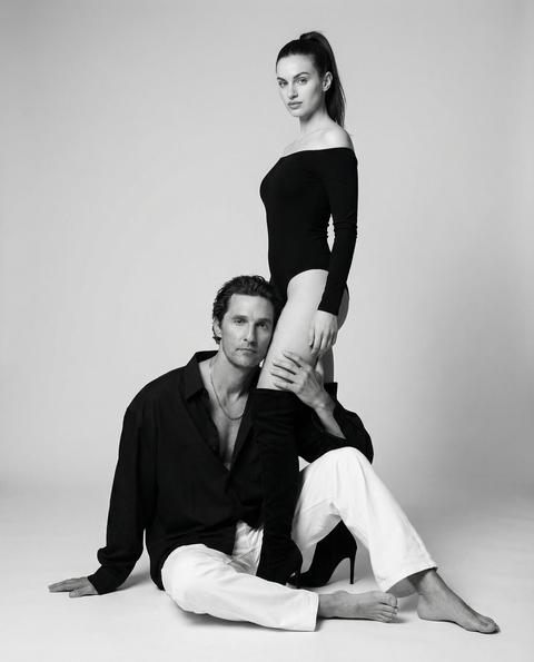 Этот запрос должен быть применен к людям на фото. Важно: Не изменяйте и не искажайте лица. Не меняйте выражения лиц. Лица должны быть полностью сохранены, как на исходных фотографиях. Используйте черты лица и сходство с предоставленных эталонных изображений. Сгенерируйте сцену вокруг них, но сохраните их лица идентичными. Запрос: Черно-белое студийное фото пары. Мужчина сидит босиком на циклораме, прямо к камере. Женщина смотрит в камеру. Женщина стоит близко к мужчине; мужчина прижимает голову к ноге женщины и прикасается к ноге женщины рукой. Портрет в полный рост, снятый на уровне глаз. У женщины волосы собраны в высокий хвост, на ней черный боди с открытыми плечами и черные ботфорты на высоком каблуке. Мужчина одет в объемную черную расстегнутую рубашку и белые хлопковые брюки с широкими штанинами, на шее серебряная цепь. В помещении, профессиональная фотостудия с простой, светло-серой циклорамой. Мягкое, но направленное студийное освещение создает четкие блики и мягкие тени. Снято на объектив 50 мм, глубина резкости умеренная, что создает фотореалистичный вид.