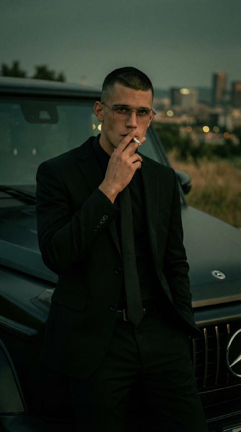 
Создай портрет, не меняя черты лица. Портрет мужчины в стиле «Old Money» и нуарной эстетике. Мужчина с короткой стрижкой «mil buzz cut» в строгом черном костюме, черной рубашке и тонком галстуке. Он в прямоугольных очках в тонкой металлической оправе, держит сигарету у рта. Поза уверенная, он опирается на капот роскошного черного автомобиля Mercedes amg 63. Сумеречное освещение, приглушенные темно-зеленые и землистые тона. Атмосфера кинематографичная, таинственная и дорогая. Глубокие тени, мягкий фокус на заднем плане с размытыми огнями города. Высокая детализация кожи и ткани, зернистость кинопленки 35мм.