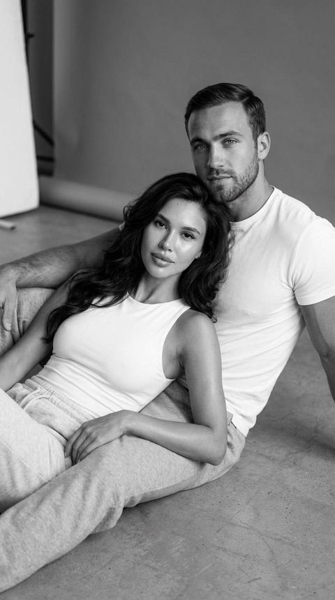 Гиперреалистичный черно-белый портрет 4K, формат 2:3, современный редакционный стиль, на котором изображена пара, лежащая на полу студии с нейтральным серым фоном. Оба смотрят прямо в камеру со спокойным и интимным выражением лица, передавая эмоциональную связь.
У женщины такое же лицо, как на эталонном изображении, с точно такими же чертами лица, красивая укладка, волнистые  распущенные волос на плечах. Выразительные глаза, натуральная кожа с легким блеском. На ней облегающий белый топ без рукавов и светло-серые хлопковые спортивные штаны. Она полулежа опирается на грудь мужчины, безмятежно отдыхая, глядя в камеру с мягким и уверенным выражением лица.
У мужчины такое же лицо, как у эталонной модели, и спокойным защитным взглядом. Подтянутая фигура. Кожа среднего тона, естественная текстура. Он носит обтягивающую белую футболку и серые спортивные штаны, его рука поддерживает женщину.
Освещение рассеянное, исходящее от бокового света под углом 45° слева, с белым отражателем для смягчения теней. Естественные и реалистичные текстуры-хлопок, мех и студийный пол.
Малая глубина резкости, центральный фокус на лицах, мягко размытый фон.
Стиль: кинематографический, эмоциональный, элегантный и интимный.