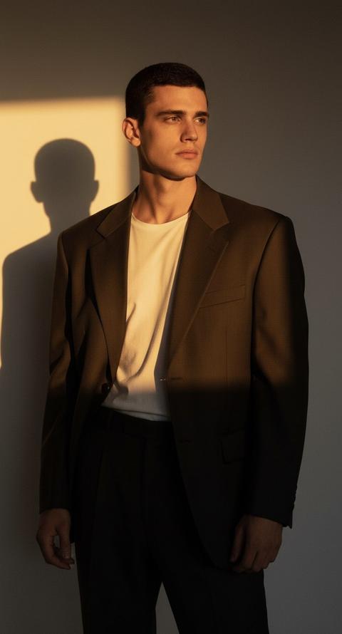 Создай портрет, не изменяй черты лица. Портрет мужчины , стоящего в драматичном контровом свете. На мужчине оверсайз  пиджак, распахнутый на груди, белая футболка. Глубокие тении жёсткие линии света на лице и шее. Золотисто-оранжевый оттенок заката (или студийного света), подчёркивающий скульптурные черты лица. Мужчина смотрит в даль с задумчивым выражением.Контровой свет, жёсткий свет, низкое солнце, золотой час, солнечные лучи, световой треугольник.тень от головы на стене, контурный свет на воротнике, высокое качество, аналоговая фотография, фотография с высоким разрешением