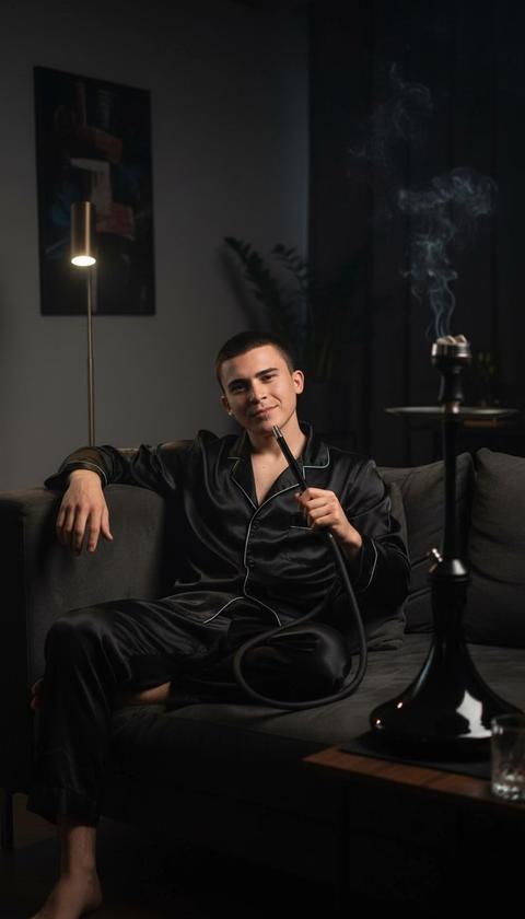 Фотореалистичный, кинематографичный снимок . Мужчина сидит в уютной гостиной, развалившись на диване. На нем мягкая пижама черного цвета. Его поза расслаблена, он смотрит прямо в камеру . Черты лица, прическа и мимика полностью сохранены и узнаваемы.
Он курит кальян, держа в руке кальянную трубку. 
Фон — темный и минималистичный интерьер гостиной. Мягкое направленное боковое освещение, словно от торшера, подчеркивает текстуру пижамной ткани и создает сложные зеркальные блики на поверхности стеклянного стакана. Тонкая контровая подсветка выделяет ее силуэт и текстуру кожи.
Технические параметры: Мелкая глубина резкости (f/1.8–2.8), объектив 85mm, низкий ISO. Высокое разрешение, реалистичные отражения и рефракция в стекле. Атмосфера — элегантная, но слегка ироничная и загадочная. Все пальцы на руках прорисованы идеально, без малейших смазываний. Не меняй черты лица