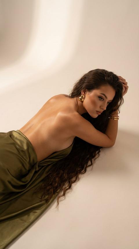 Создай:Фотография в портретной ориентации — художественный кадр с минималистичным фоном светло-бежевого/кремового цвета. В кадре находится женщина, лежащая на боку по диагонали от нижнего левого к верхнему правому углу снимка. Верхняя часть её тела — спина и бок — обнажена; нижняя часть прикрыта оливковой или темно - оливковой атласной тканью/юбкой, которая даёт мягкий блестящий перелив и образует складки вдоль бедра.
Поза расслабленная: голова оперта на согнутые руки, локти согнуты, лицо частично прикрыто рукой и направлено в сторону от камеры. Волосы очень длинные, густые и очень прямые , рассыпаны по полу . Макияж эффектный , профессиональный , загорелая кожа, акцент на скуловой зоне, ресницы, стрелки. Сходство 100%.На ушах видны круглые массивные золотые серьги. На руках золотые браслеты . Кожа модели имеет тёплый тон, на спине и плечах заметны мягкие световые блики и тонкие тени, создающие объём и подчёркивающие линию плечевого пояса и изгибы корпуса.Освещение мягкое, направленное сверху/слева, без резких контрастов — снимок выглядит атмосферным, спокойным и эстетичным. Композиция использует много пустого пространства справа и вверху, что усиливает ощущение уюта и фокус на силуэте.Высококачественный макро-портрет. Высокая детализация 4к.Не меняй черты лица. Создай:Фотография в портретной ориентации — художественный кадр с минималистичным фоном светло-бежевого/кремового цвета. В кадре находится женщина, лежащая на боку по диагонали от нижнего левого к верхнему правому углу снимка. Верхняя часть её тела — спина и бок — обнажена; нижняя часть прикрыта оливковой или темно - оливковой атласной тканью/юбкой, которая даёт мягкий блестящий перелив и образует складки вдоль бедра.
Поза расслабленная: голова оперта на согнутые руки, локти согнуты, лицо частично прикрыто рукой и направлено в сторону от камеры. Волосы очень длинные, густые и очень прямые , рассыпаны по полу . Макияж эффектный , профессиональный , загорелая кожа, акцент на скуловой зоне, ресницы, стрелки. Сходство 100%.На ушах видны круглые массивные золотые серьги. На руках золотые браслеты . Кожа модели имеет тёплый тон, на спине и плечах заметны мягкие световые блики и тонкие тени, создающие объём и подчёркивающие линию плечевого пояса и изгибы корпуса.Освещение мягкое, направленное сверху/слева, без резких контрастов — снимок выглядит атмосферным, спокойным и эстетичным. Композиция использует много пустого пространства справа и вверху, что усиливает ощущение уюта и фокус на силуэте.Высококачественный макро-портрет. Высокая детализация 4к.Не меняй черты лица.