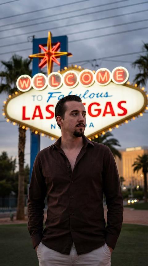 Красивый молодой человек, излучающий сдержанную уверенность, застыл в кадре. Его точеный подбородок подчеркнут тщательно выбритой темной эспаньолкой, аккуратно обрамляющей рот и подбородок с ноткой грубоватой элегантности. Его волосы, глубокого, блестящего черного цвета, безупречно зачесаны назад, каждая прядь точно уложена, мягко поблескивая в окружающем свете. Он одет в расстегнутую темно-коричневую рубашку на пуговицах; тонкая ткань — возможно, мягкий, ворсистый шелк или богатый, дышащий лен — свободно, без усилий ниспадает складками вокруг его торса, намекая на подтянутое телосложение под ней. Глубокий, землистый оттенок его рубашки создает яркий контраст с его светлыми брюками, которые плавно струятся, их ткань намекает на удобную, но изысканную посадку. Его руки небрежно, почти инстинктивно, засунуты в карманы этих светлых брюк, создавая расслабленную и тонко уверенную позу. Его взгляд, глубокий и интроспективный, пристально направлен вправо; его глаза выражают задумчивое, возможно, отстраненное размышление, легкая морщинка на лбу намекает на момент тихого осмысления.

Позади него, показанная с красивым, эфирным размытием из-за малой глубины резкости, культовая вывеска "Welcome to Fabulous Las Vegas Nevada" вспыхивает великолепным, почти осязаемым сиянием. Ее яркий неон, ослепительная мозаика рубиново-красных, сапфирово-синих и бриллиантово-золотых огней, пульсирует раскаленной жизнью, рассеиваясь в сумеречном воздухе и раскрашивая влажный сумрак своим электрическим свечением. Небо над головой — это полотно приглушенных, тяжелых, пасмурных тонов, переходящих от мягких грифельно-серых к более глубоким, синевато-пурпурным по мере полного наступления сумерек, отфильтровывая последние остатки дневного света. На фоне этого угасающего неба вырисовываются величественные пальмовые листья, их отчетливые, текстурированные формы добавляют нотку экзотики городскому пейзажу. Далее вдали неясные, мерцающие очертания зданий возвышаются подобно призрачным монументам, их края смягчены атмосферной дымкой и приближающейся ночью. Вся сцена залита мастерским кинематографическим освещением, где четкие, драматичные блики определяют контуры лица мужчины и складки его одежды, в то время как размытый фон светится почти живописным свечением, создавая захватывающую, сверхдетализированную картину, полную атмосферы.
