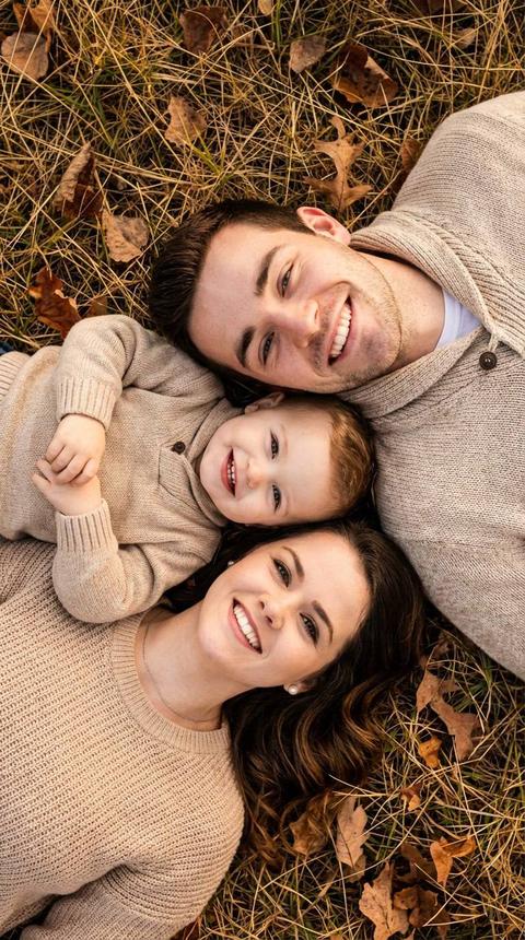 Реалистичное фото в 8к качестве семьи, лежащие на земле, образуют композицию, сфокусированную на лицах и позитивном настроении. Лица взрослых и детей смотрят в кадр, Каждый человек близко лицо к лицу, и все лежат горизонтально , относительно вертикальной фотографии. Только туловище детей повернуто в левую сторону, а мужчины и женщины в правую сторону. Дети, одеты в вязаные свитера, создавая ощущение тепла и уюта. Ребенок, широко улыбается, смотря в камеру. Дети лежат посередине между родителями. Все лежат близко друг к другу. Взрослые тоже в вязаных уютных эстетичных свитерах. Окружающая среда - сухая трава и редкие опавшие листья, указывающие на осенний сезон.  Общая тональность -теплая, с преобладанием нейтральных оттенков коричневого и бежевого, создающая расслабленную и естественную атмосферу. Ракурс – сверху, с небольшой глубиной резкости.  Фотография вызывает ощущение семейной теплоты и спокойствия. Сохрани черты лица всех людей изображенных на референсе максимально точно, не искажая их