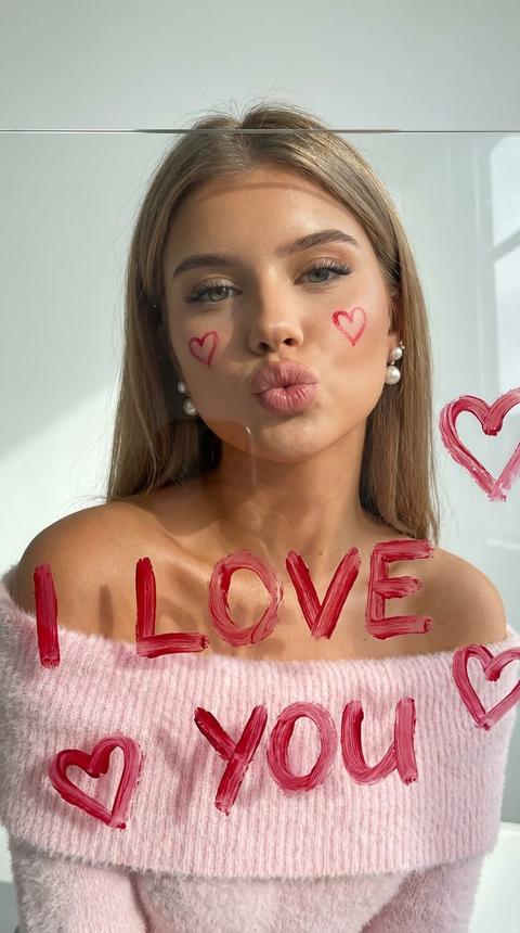 Создай портрет, не меняя черты лица. Крупный план женщины  с длинными  волосами, которая смотрит сквозь прозрачное стекло. Она сложила губы для поцелуя. На щеках нарисованы два маленьких красных сердечка. На переднем плане, прямо на стекле, ярко-красной помадой или краской небрежно написаны слова «I LOVE YOU» и нарисованы сердечки. Мазки краски объемные и текстурные. Она одета в нежно розовый объемный пушистый свитер, который спадает с плеча, на ушах жемчужные серьги, безупречный макияж, кожа слегка загорелая . Мягкое естественное освещение, светлый минималистичный фон, теплая цветовая гамма. Высокая детализация кожи и текстуры свитера.
