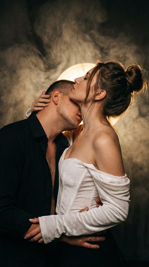 Кинематографический стиль с добавлением зерна
Крупный план лиц мужчины и женщины
Она в белой корсетной бархатной кофте с рукавами частично кофта спущена с плеча 
Мужчина в растегнутой чёрной рубашке  
За ними светит тускло светлый свет 
Между ними страсть мужчина обнимает её и целует в шею 
Волосы у девушки собраны в небрежный пучок, локоны выбиваются
В камеру не смотрят  
Тусклый свет 
Дым вокруг. Сохрани черты лица, не изменяя их. 