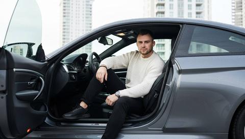 Реалистичное фото мужчины, примерно 30 лет, сидит в салоне спортивного автомобиля BMW m4 G82. На нем белый свитер крупной вязки, черные брюки, черные кроссовки , часы на запястье и браслет. Мужчина слегка откинулся, скрестив ноги, рука расслабленная лежит на колене одной ноги , вторая нога стоит ровно, колено направлено в камеру. его взгляд направлен в камеру, выражение лица спокойное, серьезное , но с легкой игривостью. Положение тела естественное. Автомобиль цвета мокрый асфальт, дверь открыта, создавая ощущение простора. Салон кожаный. За спиной мужчины, через открытую дверь, виден современный белый архитектурный фон с высокими зданиями, отражающими солнечный свет. Освещение мягкое, естественное, подчеркивающее фактуру материалов. Общая атмосфера - современная, стильная, с ноткой расслабленности и уверенности. Снимок выполнен в стиле современной фотографии, с акцентом на детали и текстуры. Сохрани максимально черты лица мужчины, не искажая их.